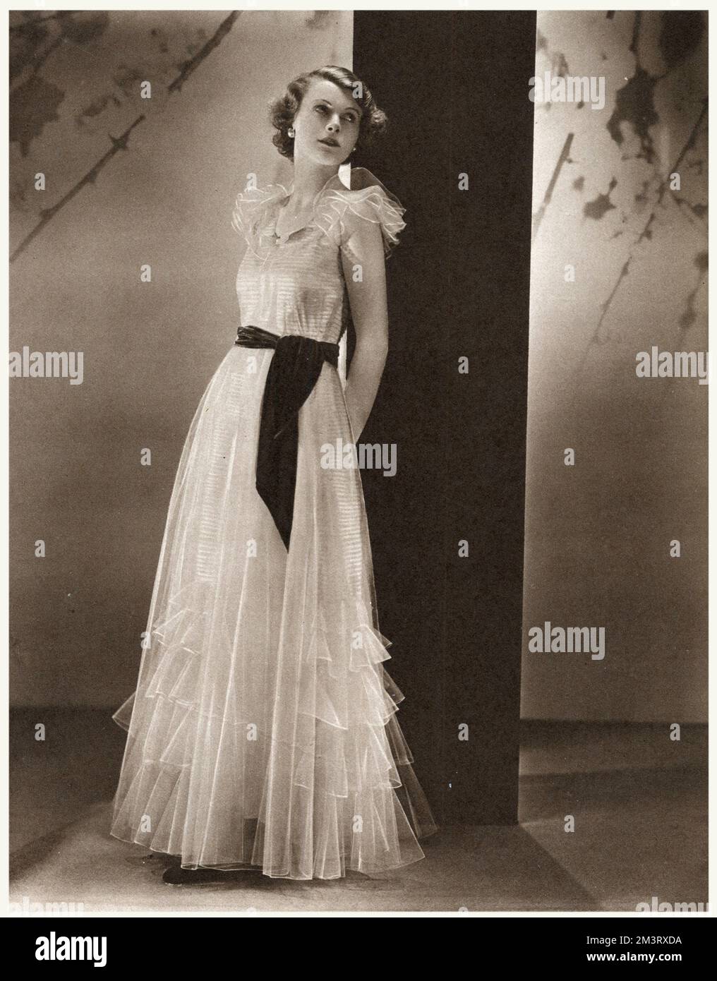 A debutante frock in primrose yellow net with a rich brown velvet sash.  Delicately tucked panels, ending in ever-widening frills, give an air of gossamer lightness to the skirt, while the shoulder frills are delightfully youthful and pretty.       Date: 1933 Stock Photo