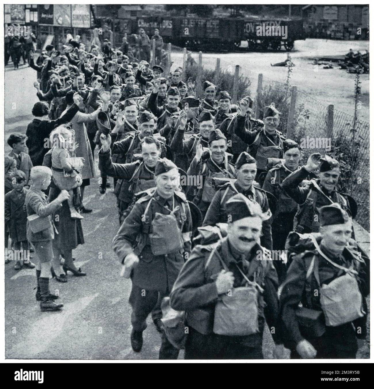 British soldiers photographed smiling as they leave their home for the Western Front, a few weeks into the Second World War. Women and children are shown waving as the men march through, some of whom raise their hats in salute.      Date: 1939 Stock Photo