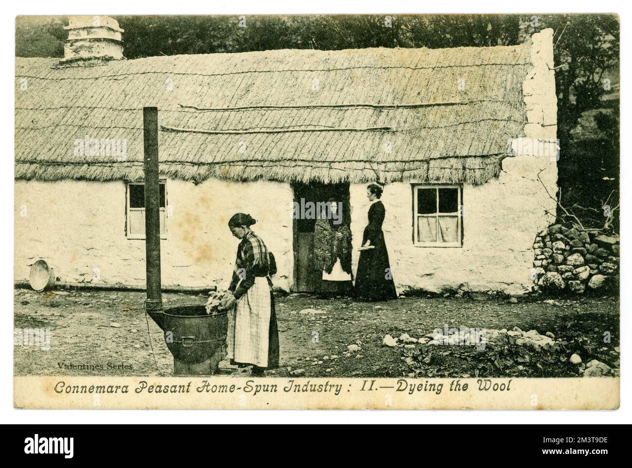 Original Edwardian era greetings postcard of peasant home-spun industry. Dyeing the Wool. outside a rustic cottage home, vernacular architecture built of local stone and roofing of reeds. Connemara, Galway, Republic of Ireland. Circa 1905. Stock Photo