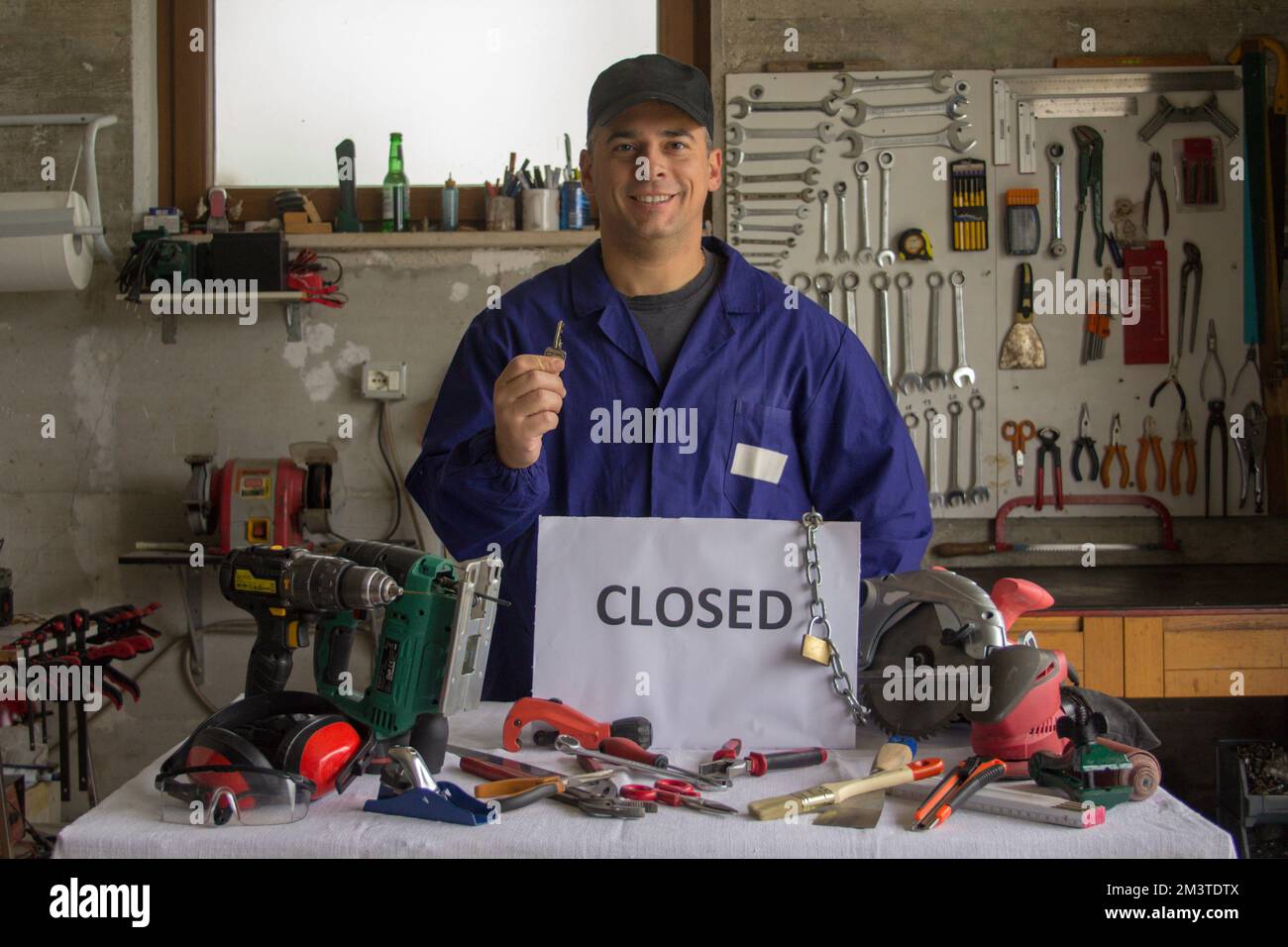 Image of a smiling craftsman in his workshop holding a wrench in his hand and in front of a bench with tools and a closed sign. Reopening of business. Stock Photo