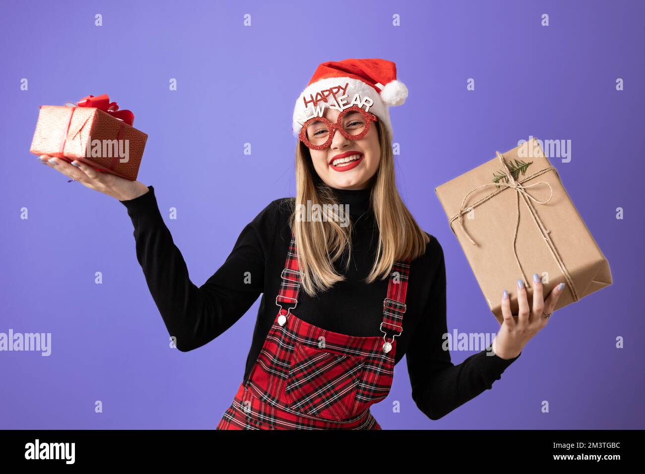 Beautiful, young, blonde girl with Santa hat and prop glasses, holding a gift boxes, bigger and smaller. Copy space Stock Photo