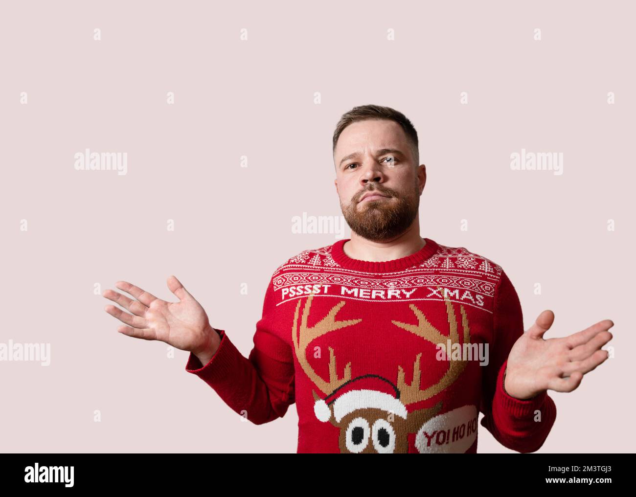 Young, bearded man shrugging shoulders in Christmas sweater, isolated on red background. Copy space Stock Photo