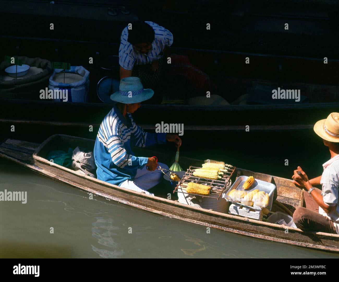 JOVENES ASAN MAZORCAS A LA PARILLA EN SU BARCA PARA VENDERLAS. Location: MERCADO FLOTANTE. BANGKOK. TAILANDIA. Stock Photo