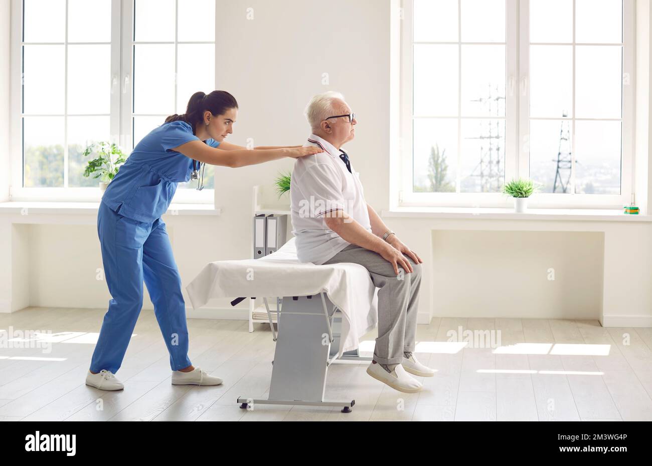 Senior man with back pain sitting on massage table while physiotherapist examines him Stock