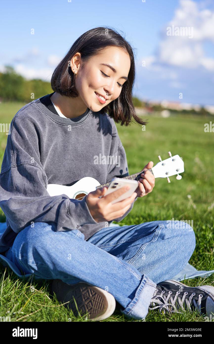 vertical shot of smiling asian girl with smartphone playing ukulele reading chords or lyrics while singing relaxing outdoors lifestyle and people 2M3WG9E
