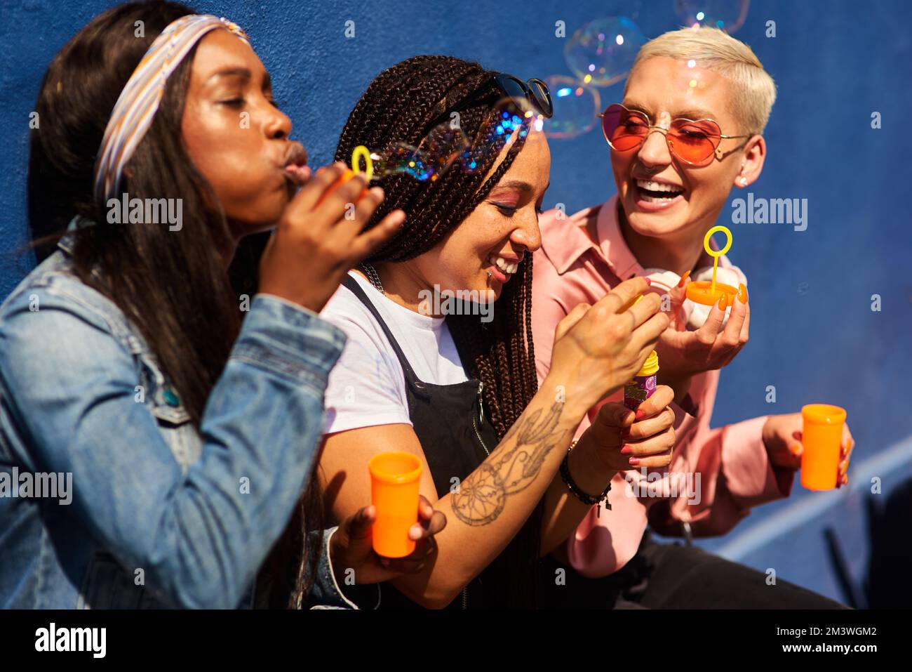 Youre never too old to blow bubbles. three attractive young women sitting against a blue wall together and bonding by blowing bubbles. Stock Photo