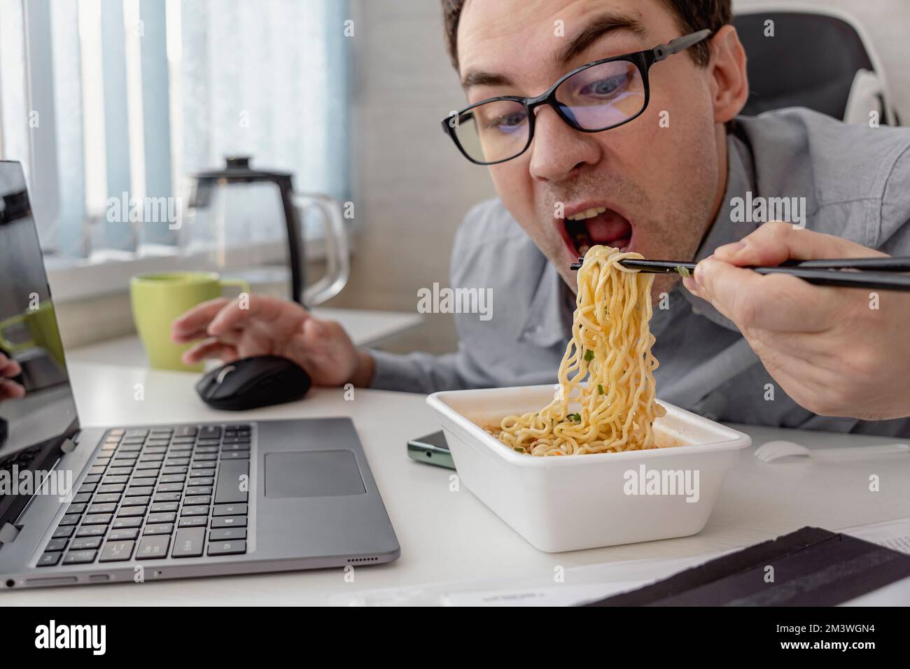 Young man eating instant noodles while working with laptop in office Stock Photo