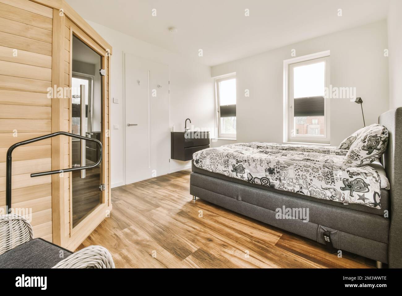 a bed in a room with wood flooring and white walls behind the bed is a large window that looks out to the outside Stock Photo