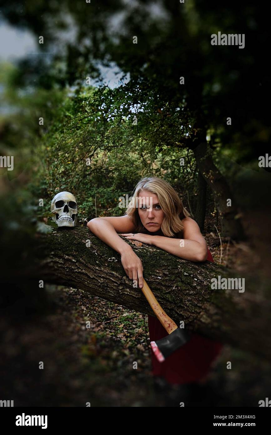 blond woman in red dress with bloodstained axe leaning on a tree with a skull, horror ,thriller, Halloween  theme Stock Photo