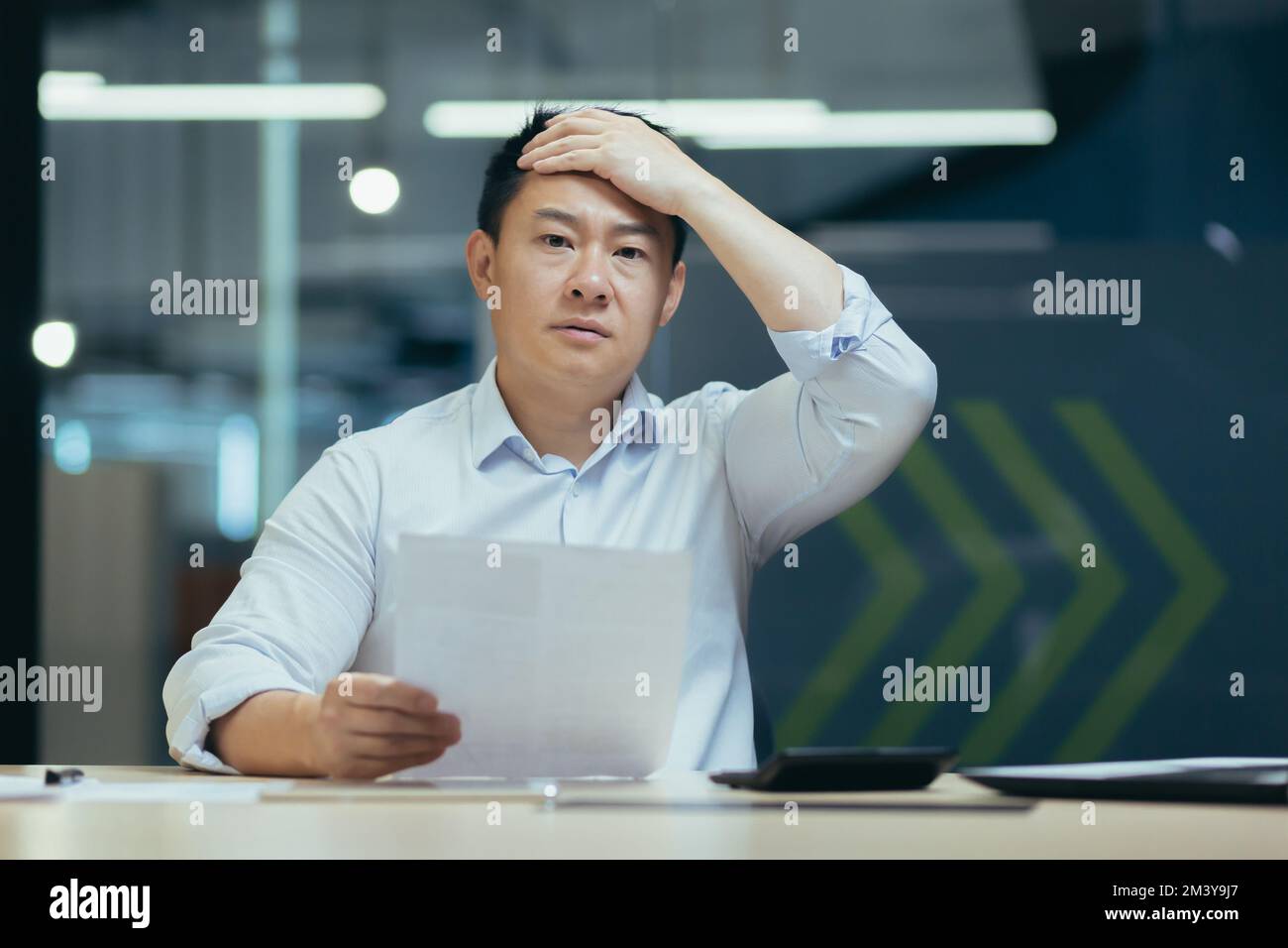 Shocked male Asian student holds a document in his hands. The worried person holds his head. Sitting at the desk in the office, received a letter, exam results, information about studies, bill. Stock Photo
