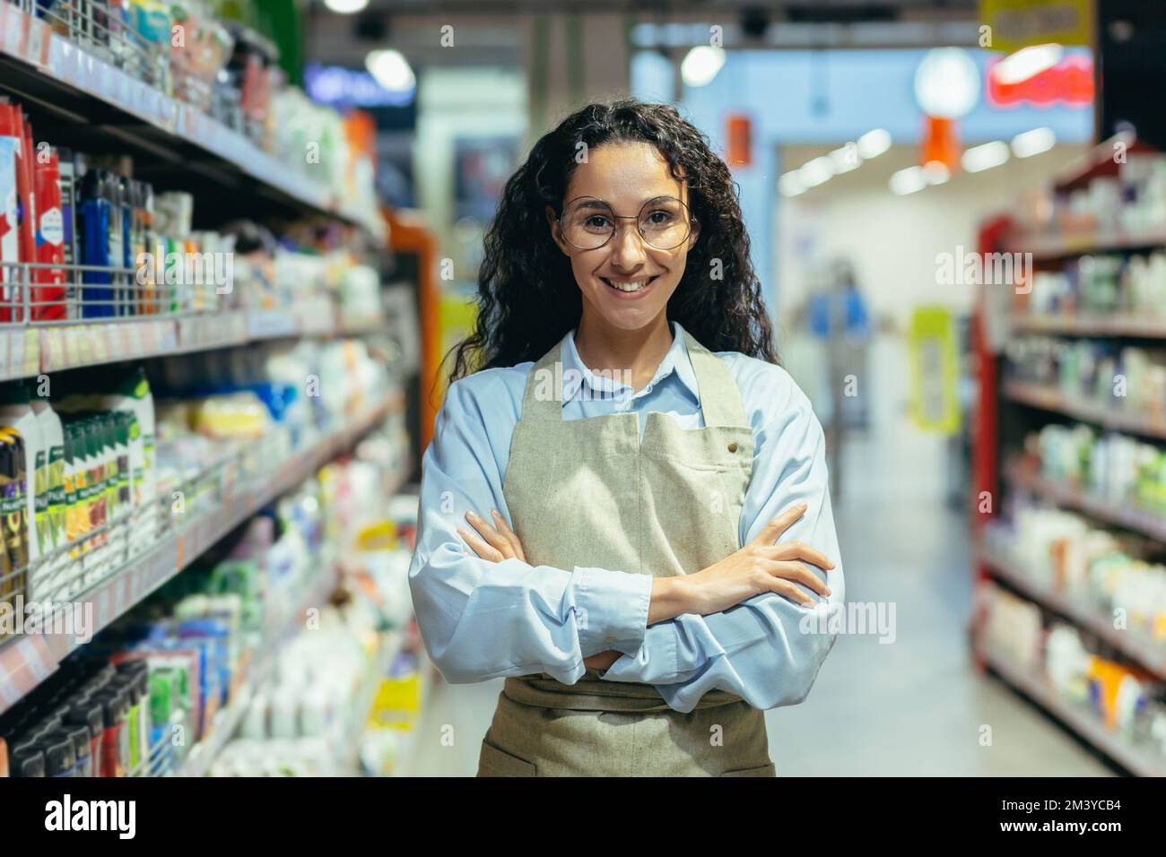 Portrait of Hispanic woman seller in supermarket, female worker in apron with curly hair and glasses smiling and looking at camera, saleswoman among goods with household chemicals and shelves. Stock Photo