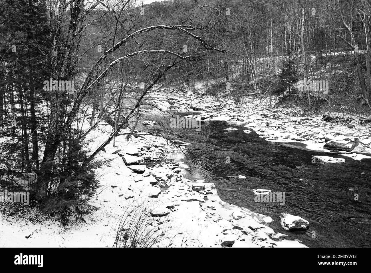 A river runs through a rural area in Vermont in mid-December surrounded by leafless trees and snow covered rocks Stock Photo