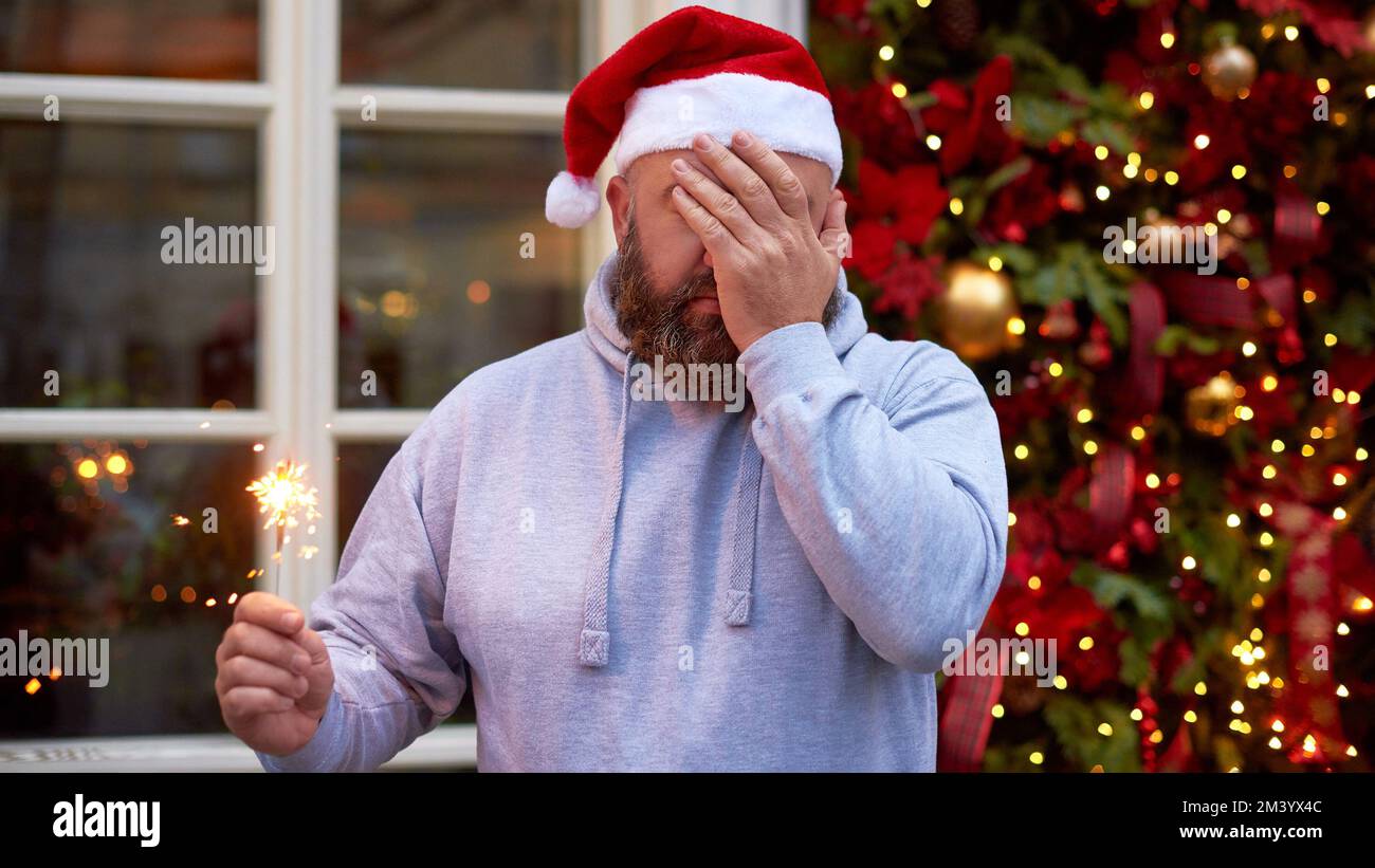 man in santa hat do facepalm gesture holding Christmas sparklers on decorated christmas tree background Stock Photo