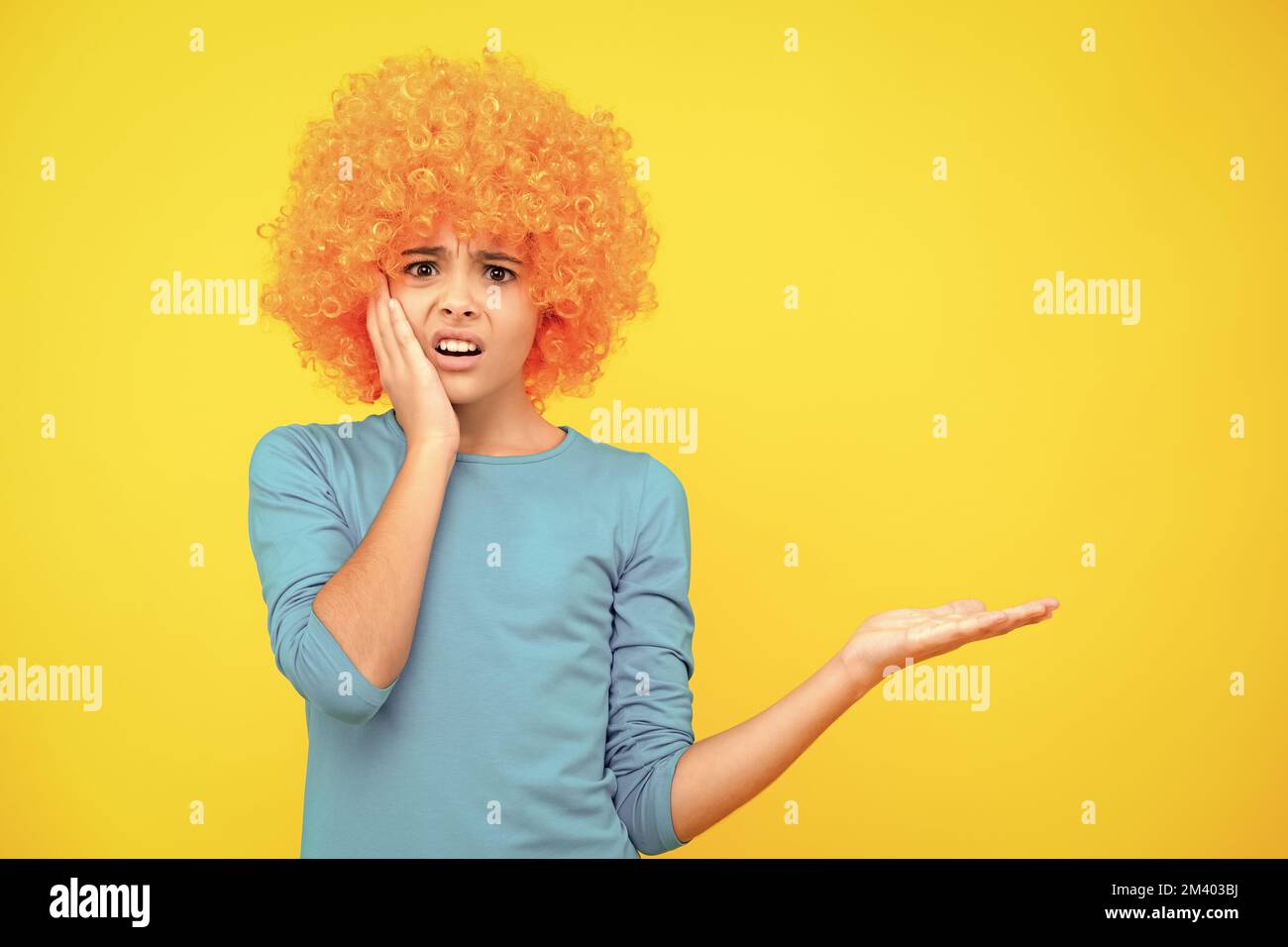 Funny kid in curly redhead wig. Time to have fun. Teen girl with orange hair, being a clown. Angry face, upset emotions of teenager girl. Stock Photo