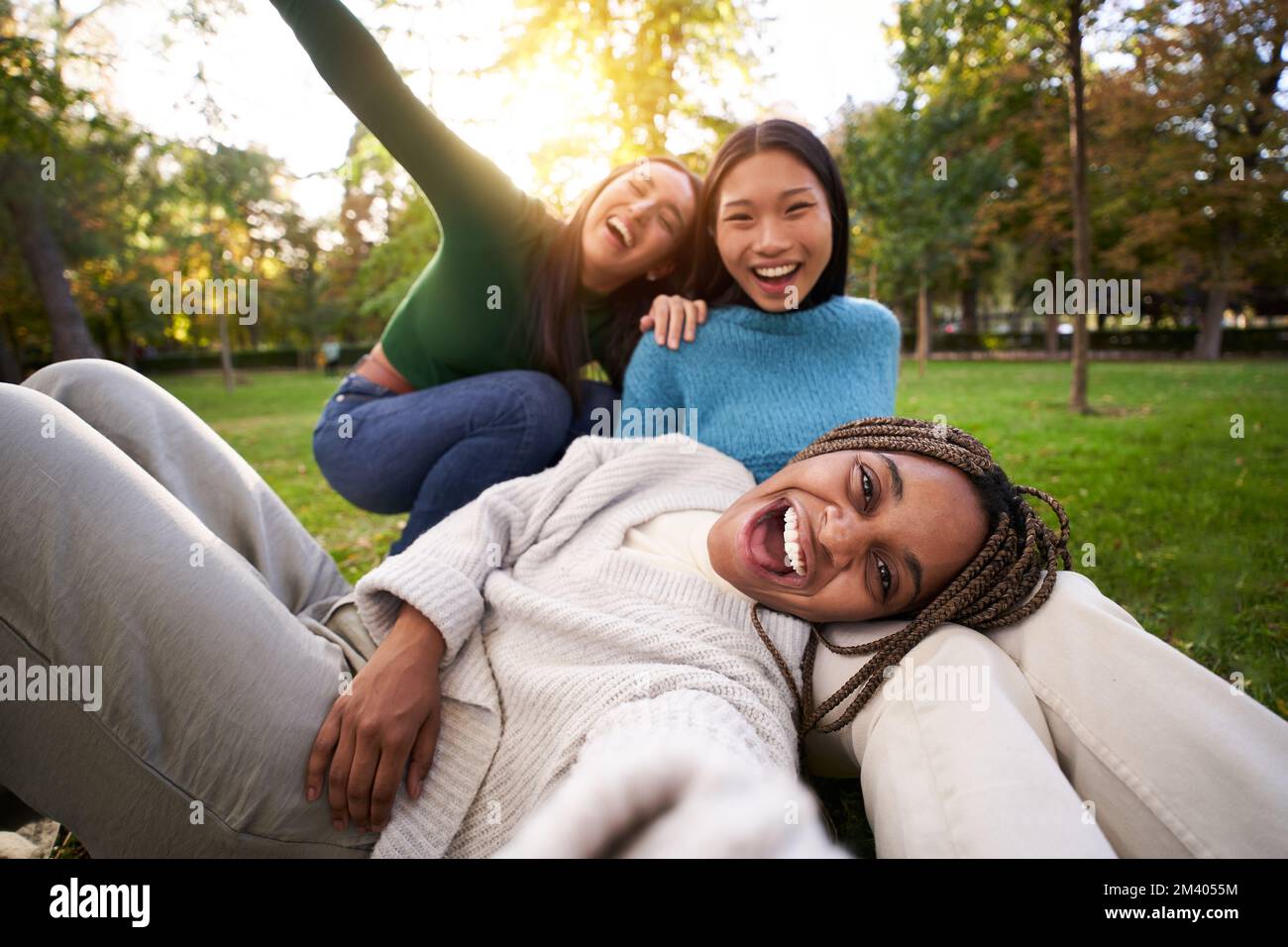 Three multiracial girls lying on the grass taking selfie looking at camera. Group of female friends Stock Photo
