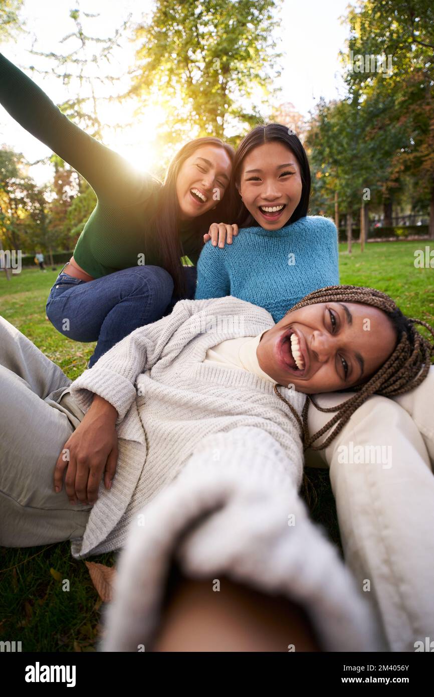 Vertical photo of Three multiracial girls lying on the grass taking selfie looking at camera. Stock Photo