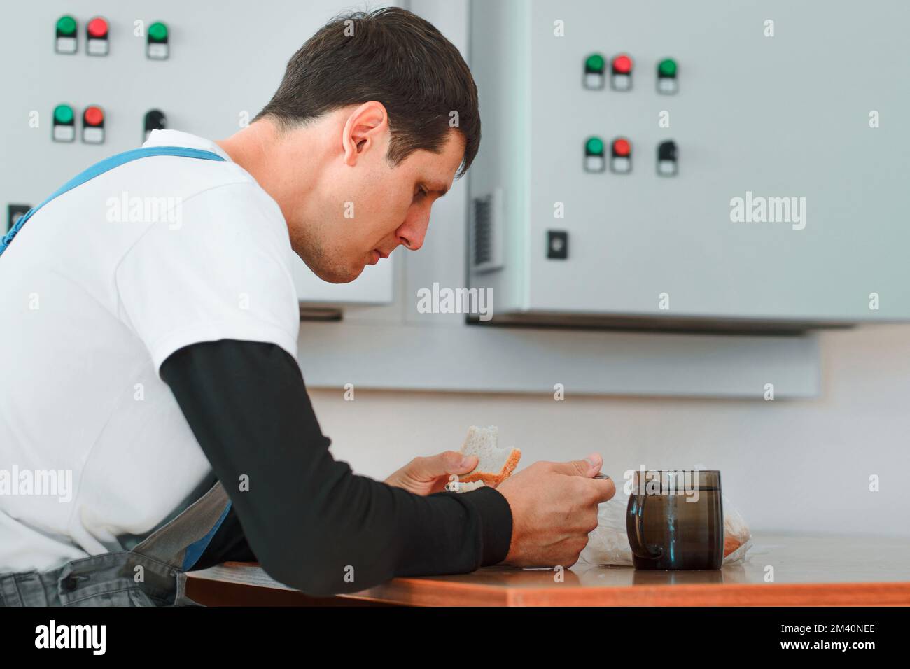 Lunch break. Eating in workplace during work. Caucasian man in work suit sits at table in production hall and eats from container. Worker has lunch in break room.. Stock Photo