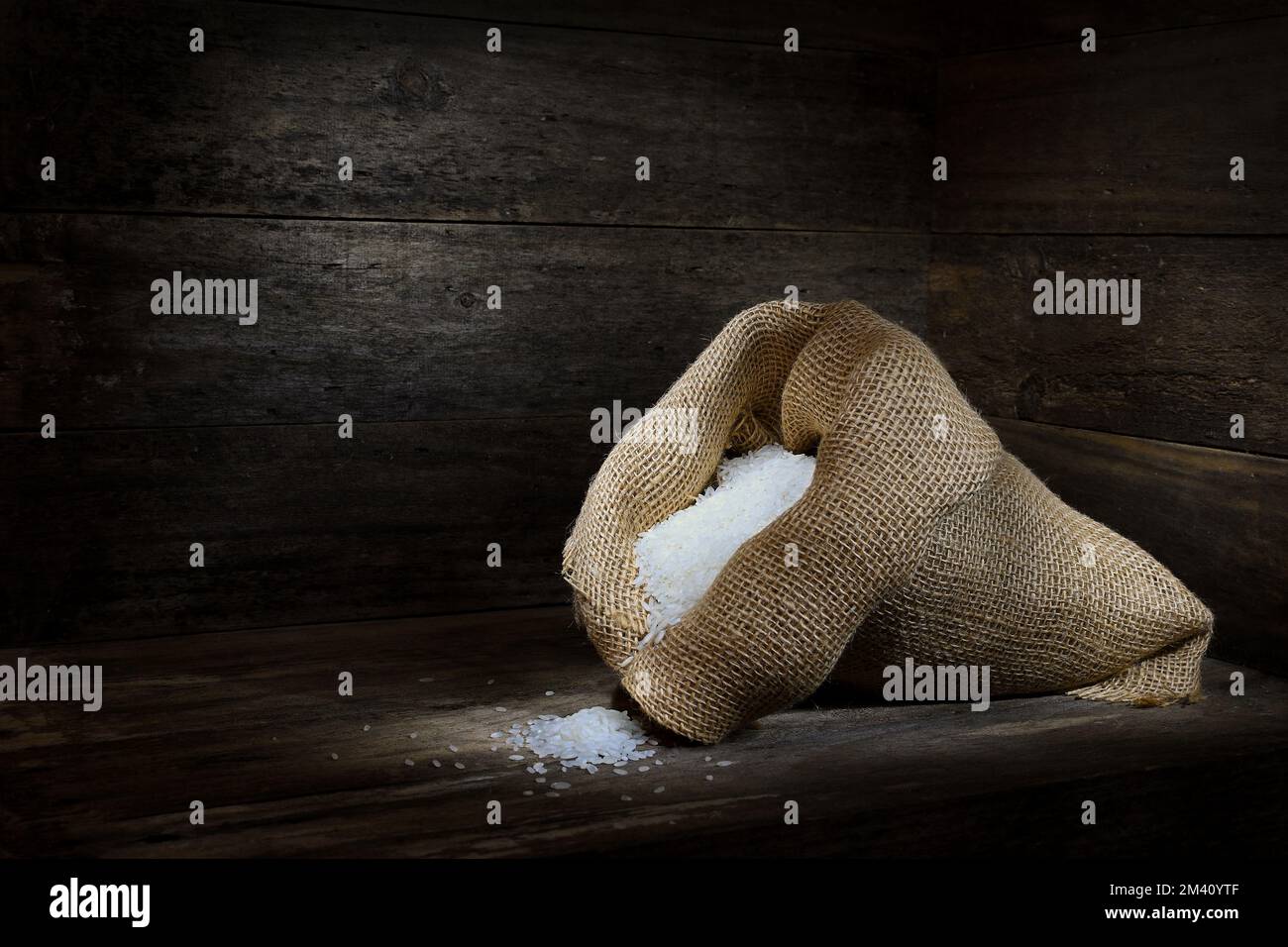 A rustic scene of a hessian bag overflowing with long grain White Rice on a wooden box-like surrounding in dark mood lighting; captured in a Studio Stock Photo