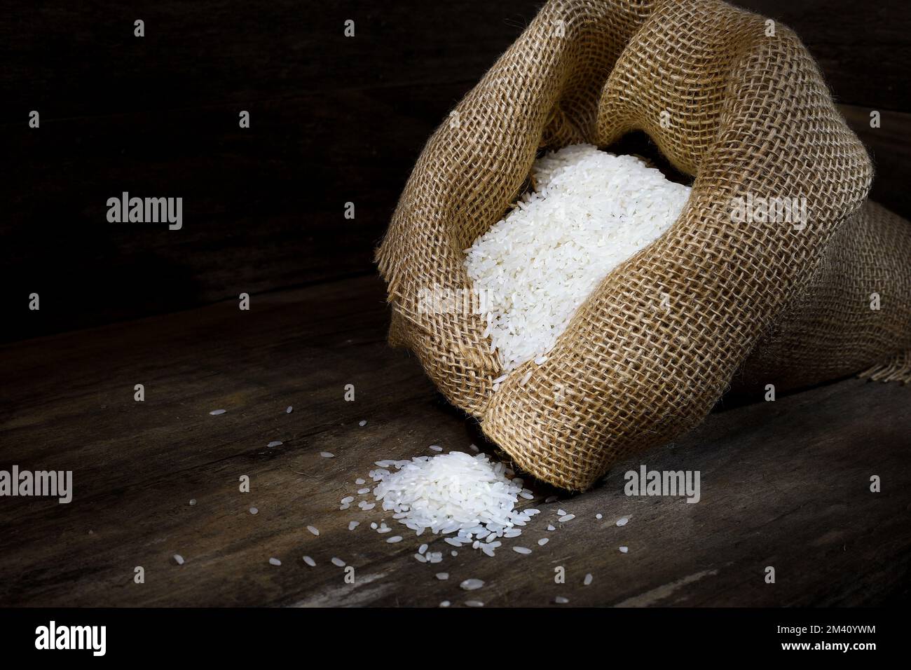 A rustic scene of a hessian bag overflowing with long grain White Rice on a wooden box-like surrounding in dark mood lighting; captured in a Studio Stock Photo