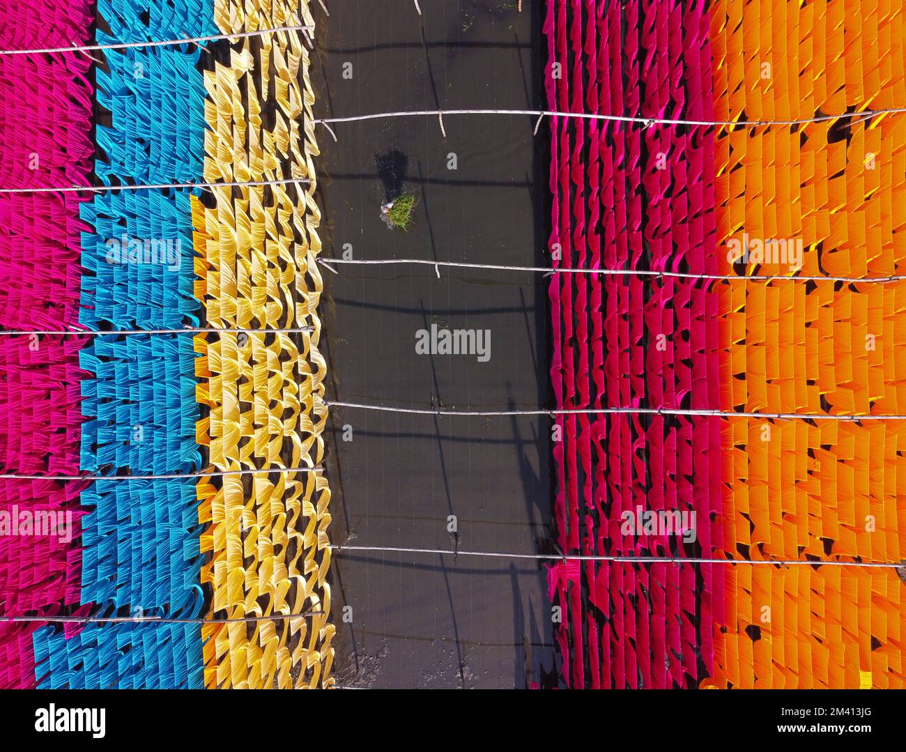Narayanganj, Dhaka, Bangladesh. 18th Dec, 2022. A dazzling network of interlocking colors produces a rainbow of colors as colorful fabrics are hung to dry above a flooded field in Narayanganj, Bangladesh. Iron wires are used between a bamboo framework to create giant washing lines for the final part of the dying process after which the cloths are made into t-shirts and vests at the garment factory. About 4,000 pieces of fabric are hung to dry here every day. The process usually takes five hours, with each set of 80 pieces at a time. Credit: ZUMA Press, Inc./Alamy Live News Stock Photo