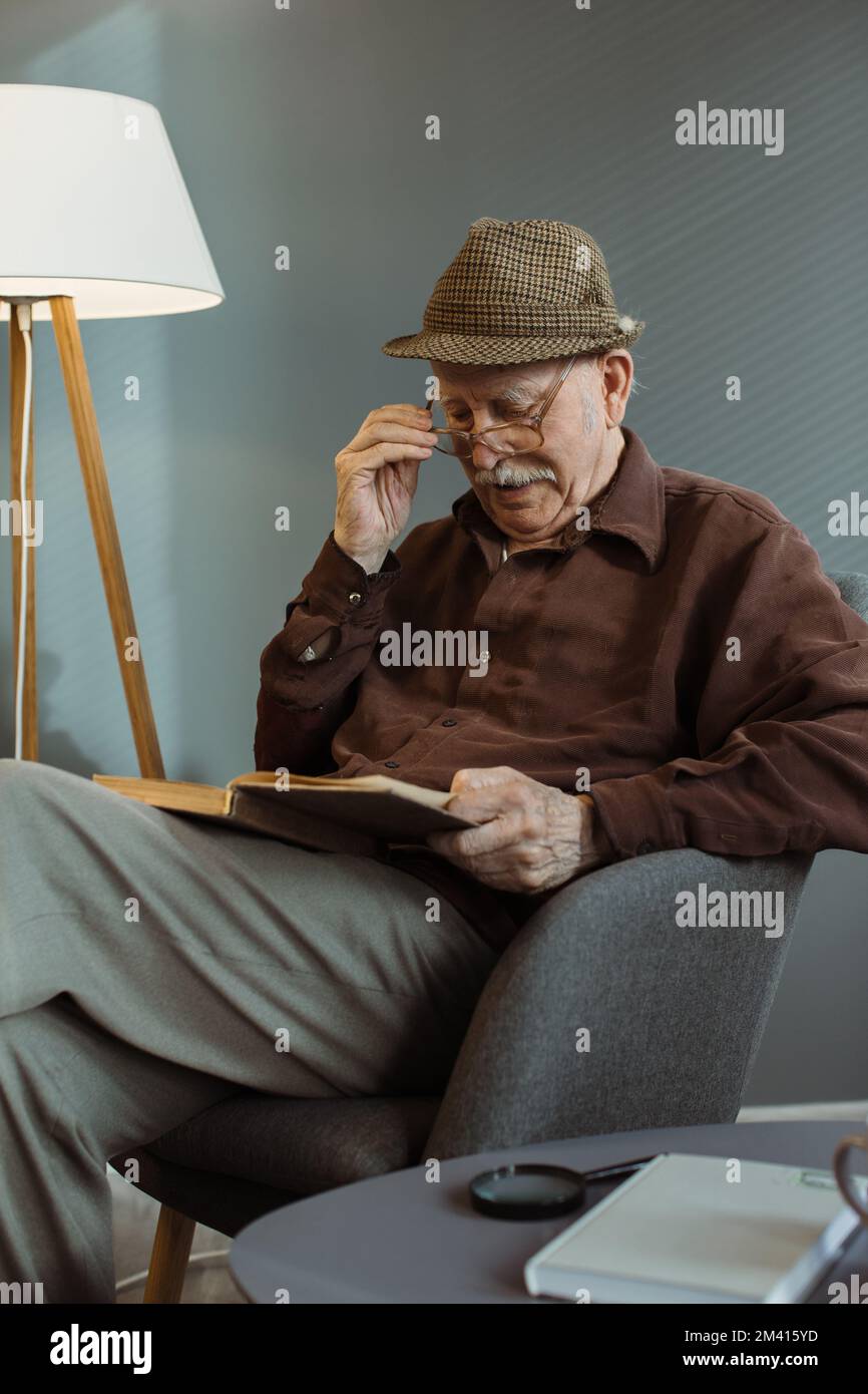 An elderly man is sitting alone on a chair in the living room, wearing glasses and reading a book. Stock Photo