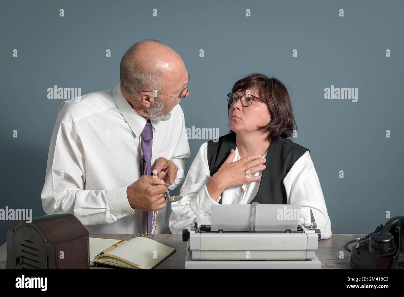 Time is up! Retro office workplace. Elderly boss talking to his expirienced secretary woman pointing finger to watch. Manual typewriter on the table. Stock Photo
