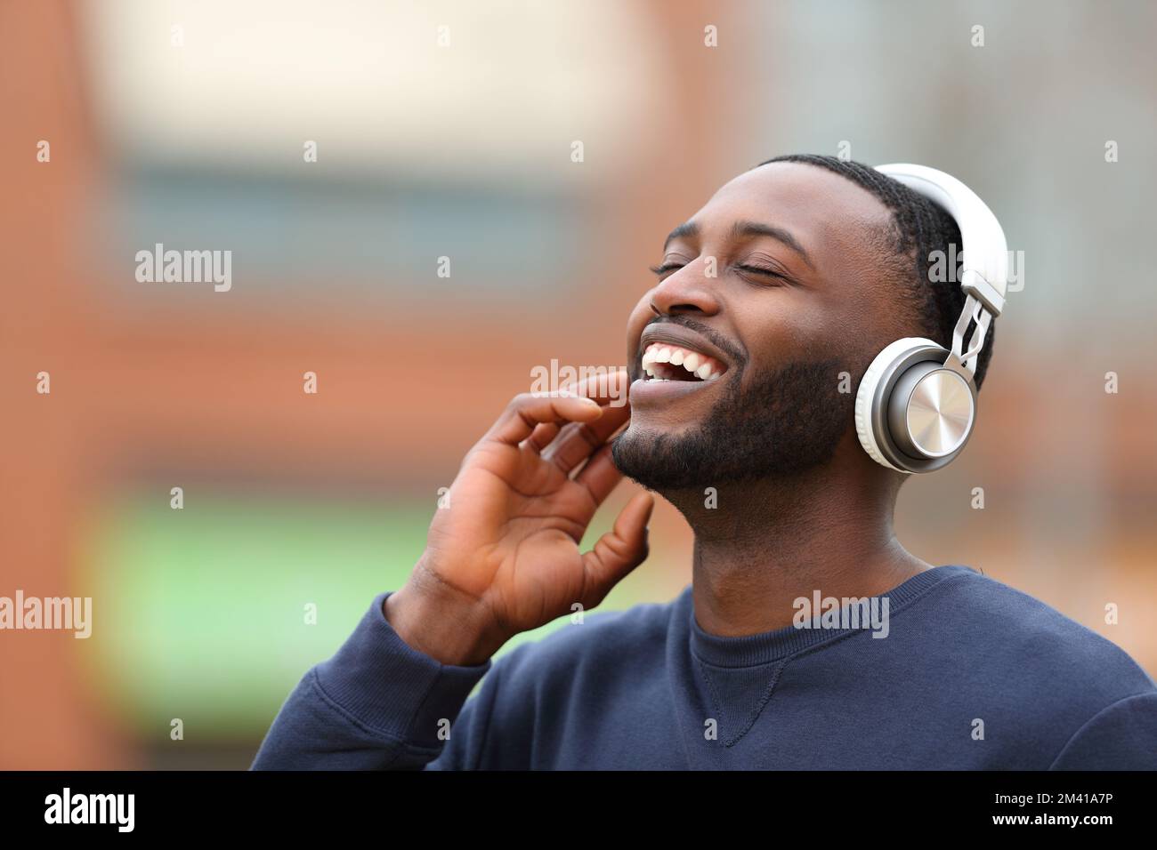 Happy black man wearing wireless headphones listening to music laughing in the street Stock Photo