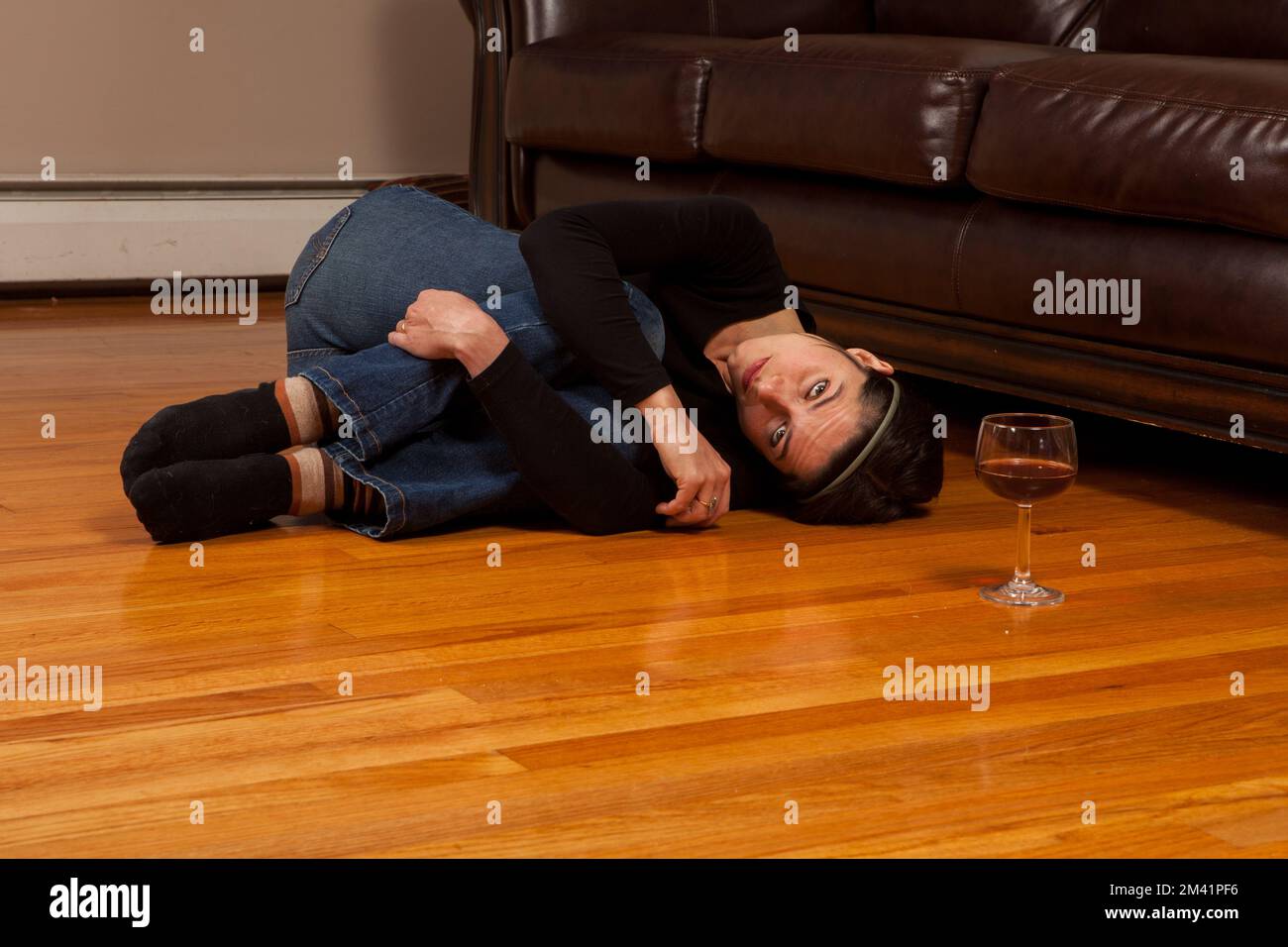 Young woman lying on the floor  in the fetal position with a wine glass looking at the camera Stock Photo