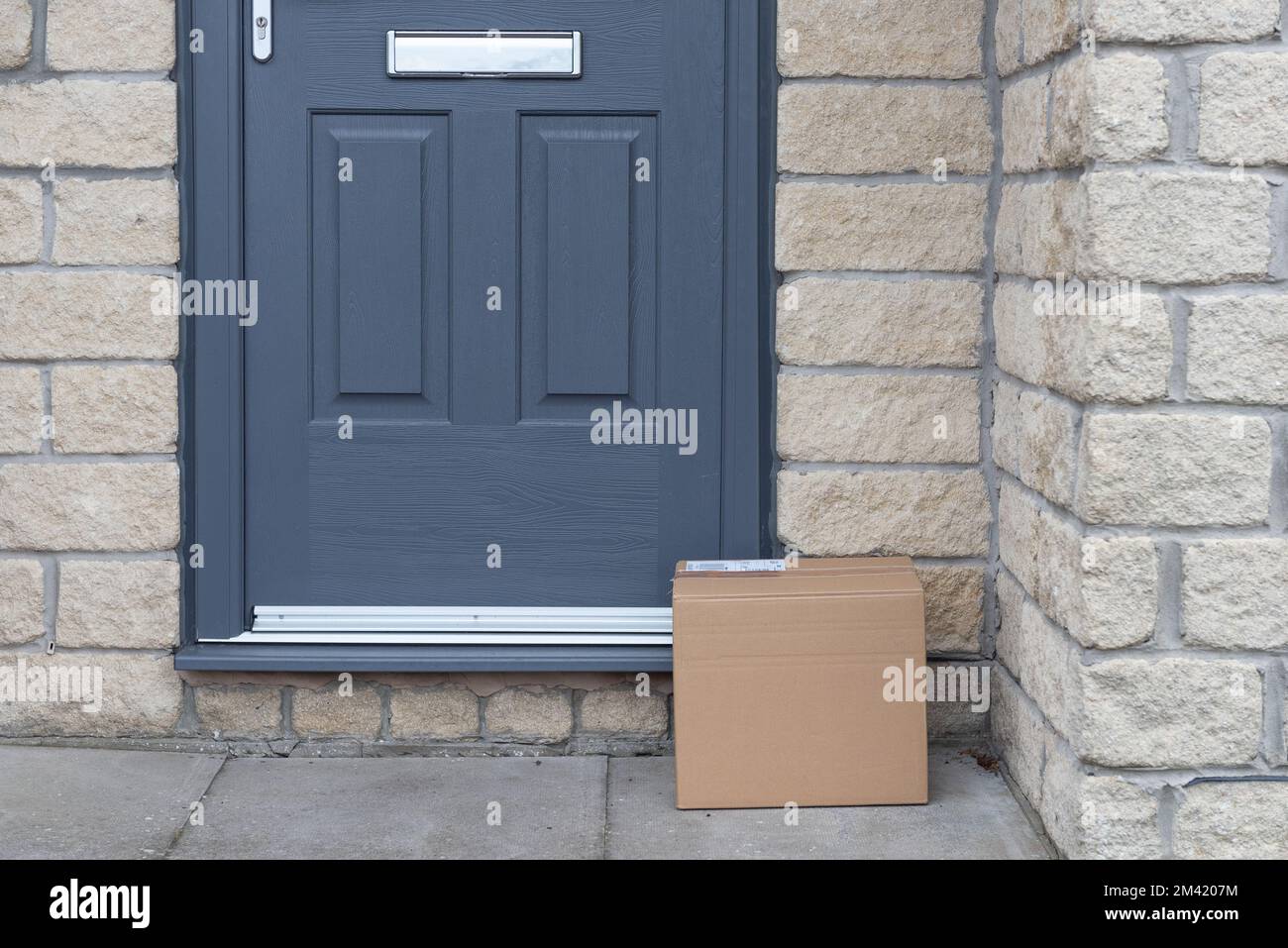 Small cardboard parcel left on the doorstep. Grey door and doorway. Courier delivery parcel. Stock Photo