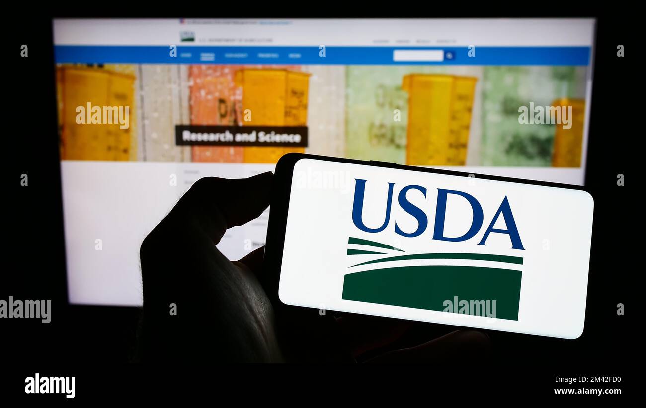 Person holding cellphone with logo of United States Department of Agriculture (USDA) on screen in front of webpage. Focus on phone display. Stock Photo