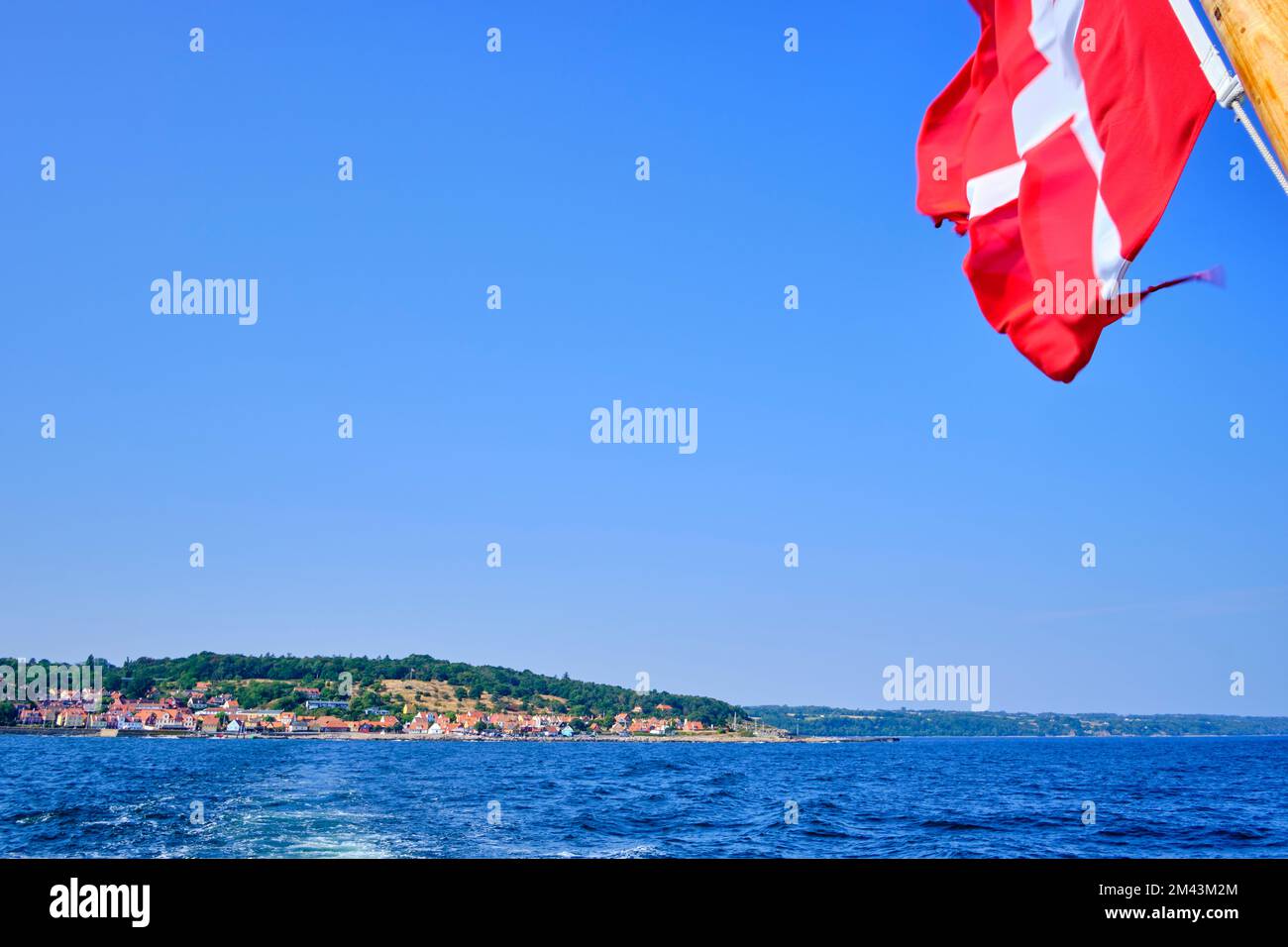 Flying Danish flag at the stern of an excursion boat and picturesque view of the coastline of Gudhjem, Bornholm island, Denmark. Stock Photo