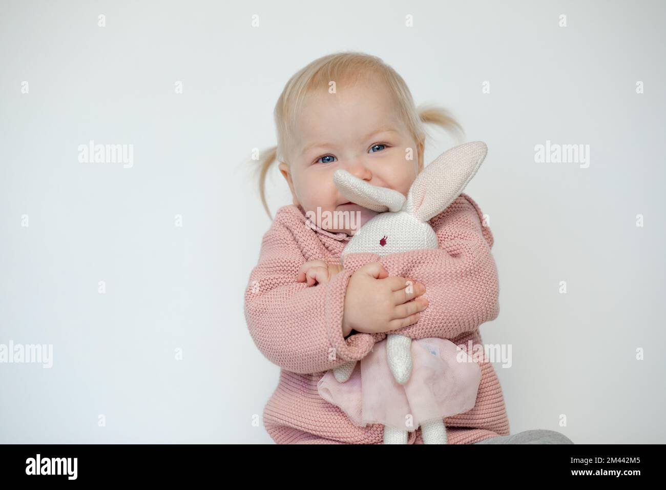 Beautiful baby girl play with stuffed animal, isolated on white. Joyous toddler happily embracing teddy bunny. Blonde haired child in pink clothes Stock Photo