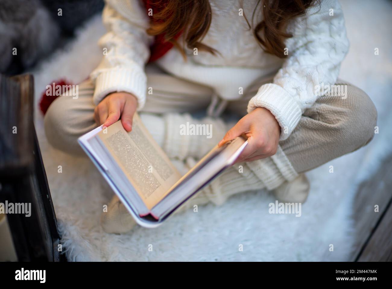 A girl with a book is sitting on the floor on a furry white carpet. Dressed in a white sweater and light trousers with a red scarf. Stock Photo