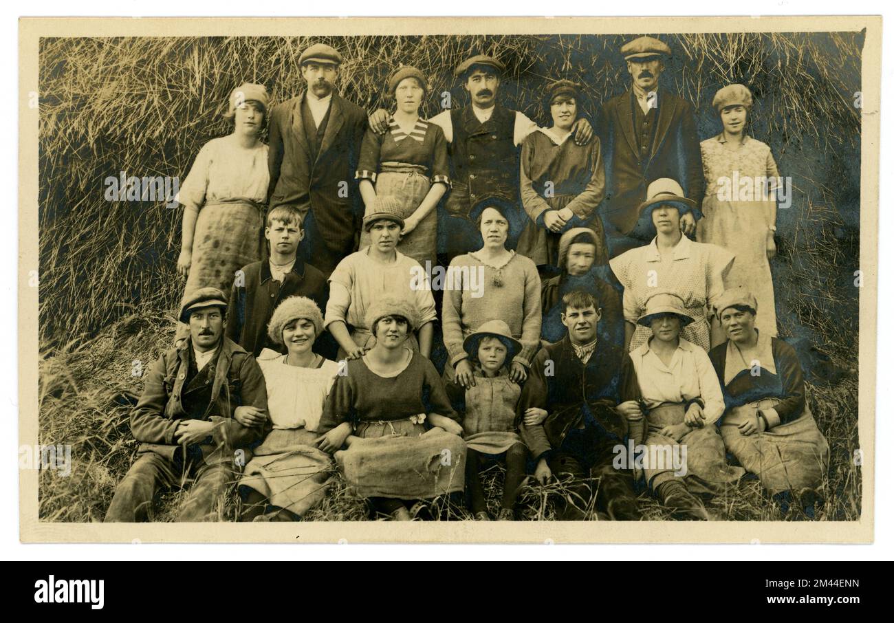 Original and charming late 1920's early 1930's era postcard of cheerful but tired looking working class labourers relaxing for a group photograph after the harvest has been brought in, big haystack behind them, lots of characters, young men and women, some children, family members. The workers' clothes are protected by sack cloth aprons. Photograph by Ashby Swift, South Street, Bourne, South Lincolnshire, England, U.K. Stock Photo