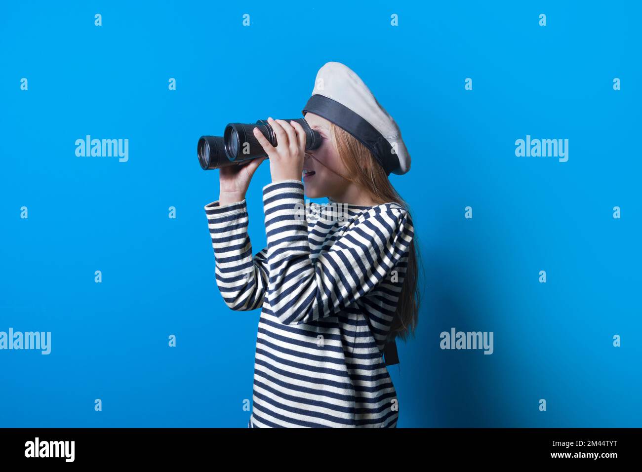 A teenage girl in a sailor suit with a binocular looking into the distance, Horizontal portrait of a sailor girl on a blue background Stock Photo
