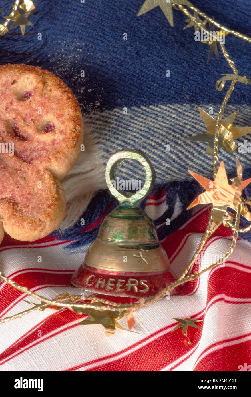 CHEERS bell surrounded by gingerbread cookie, gold stars, red and white striped fabric and blue scarf background. Closeup of celebration party time. Stock Photo