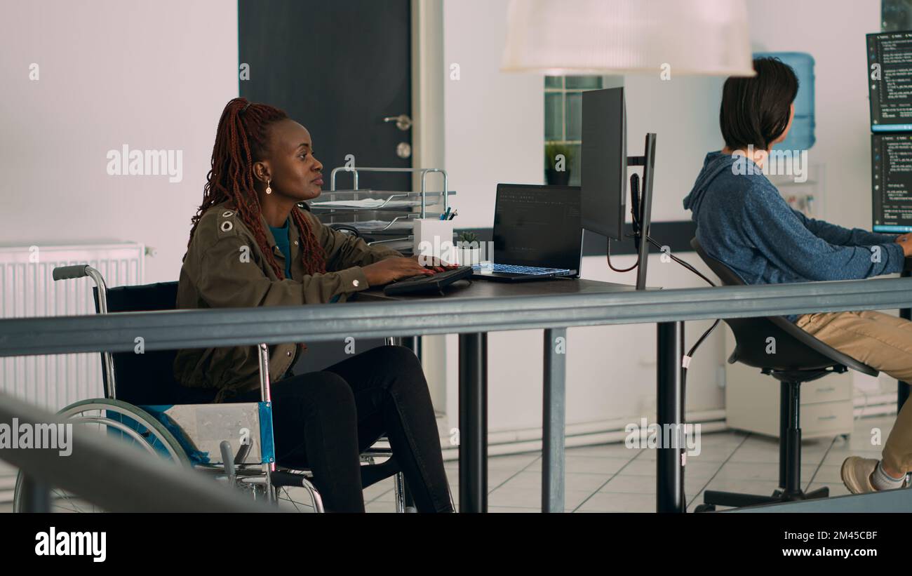 Cloud programmer in wheelchair using server data to create software app on computer, suffering from chronic disability. Coder working on new use rinterface development in agency office. Stock Photo