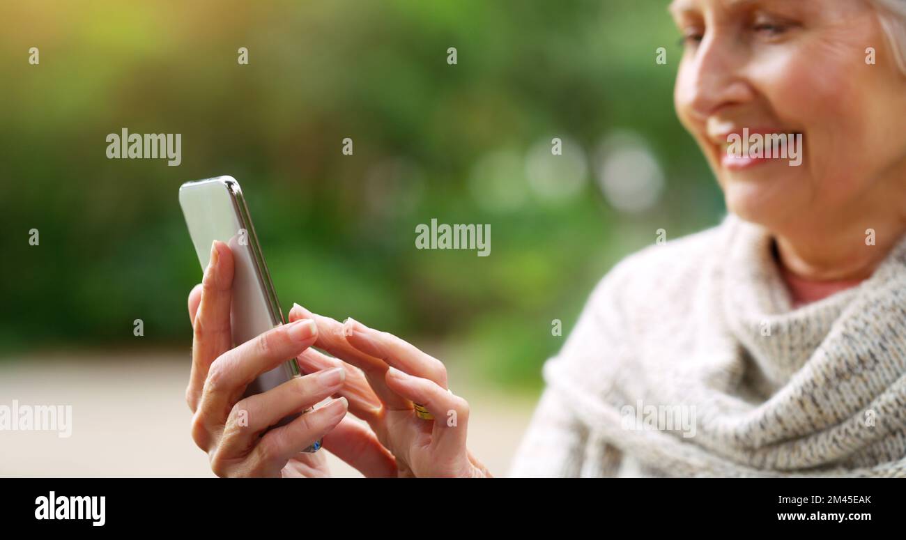 I need my reading glasses. a cheerful elderly woman texting on her cellphone while being seated on a bench outside in a park. Stock Photo