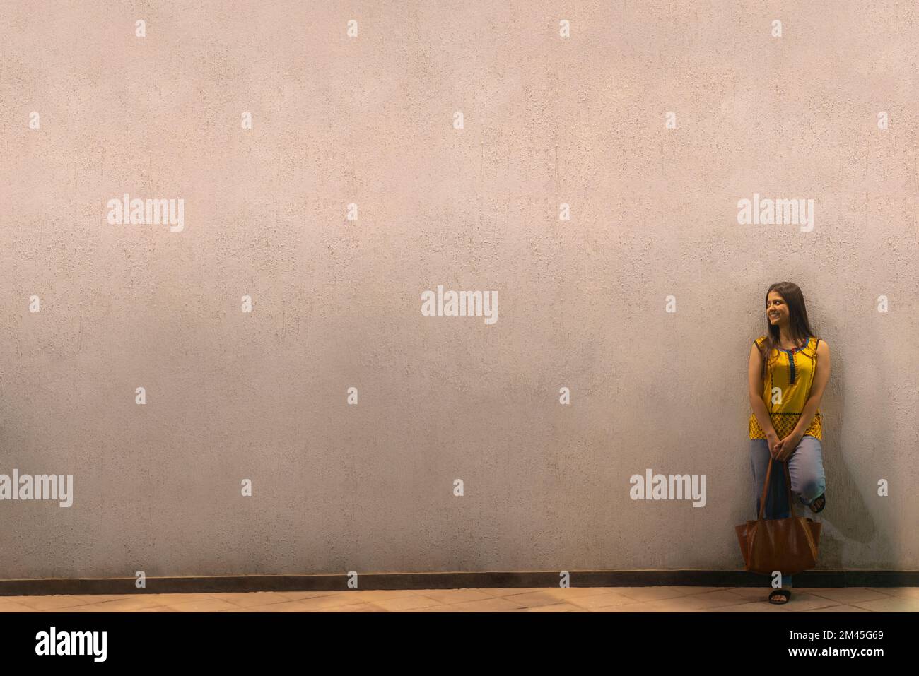 Long distance shot of an Indian woman with bag standing against wall Stock Photo