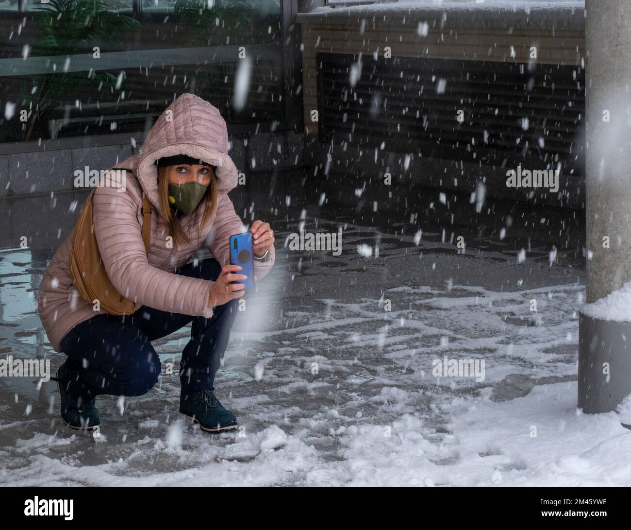 Woman wearing a face mask and holding a mobile phone while taking photos of the falling snow outdoors. Technology and winter concept. Stock Photo