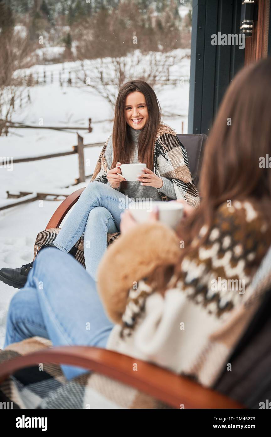 Young women enjoying winter weekends on terrace of contemporary barn house in the mountains. Two girls sitting on chairs and drinking hot tea. Stock Photo