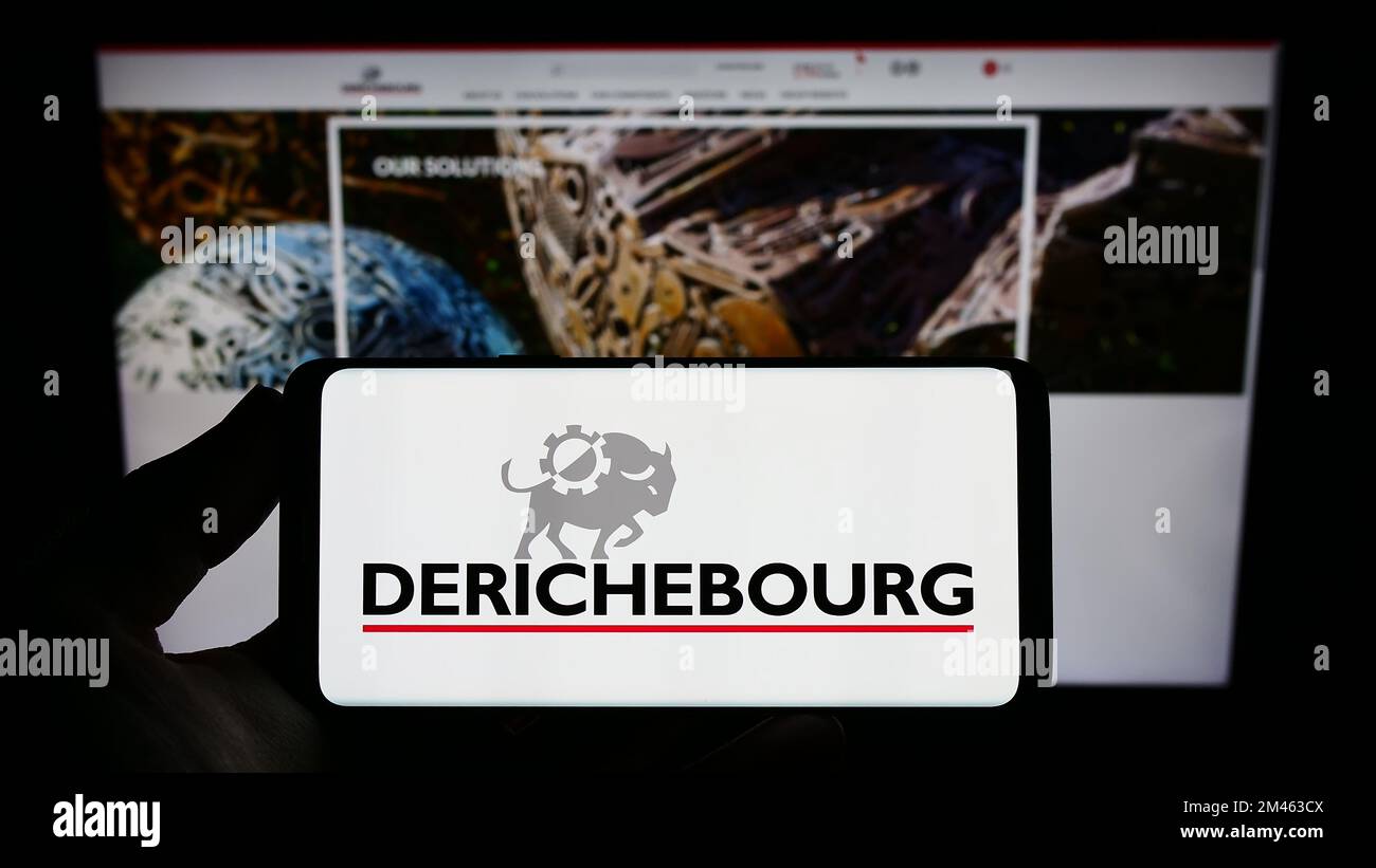 Person holding smartphone with logo of French recycling company Derichebourg S.A. on screen in front of website. Focus on phone display. Stock Photo