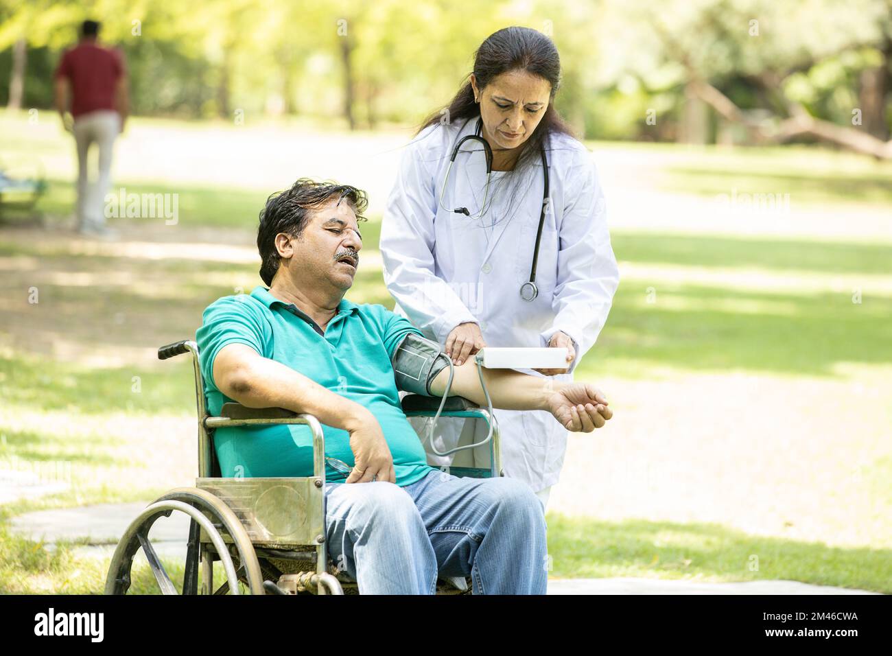 Indian doctor check blood pressure of senior male patient in a wheelchair outdoor at park. Stock Photo