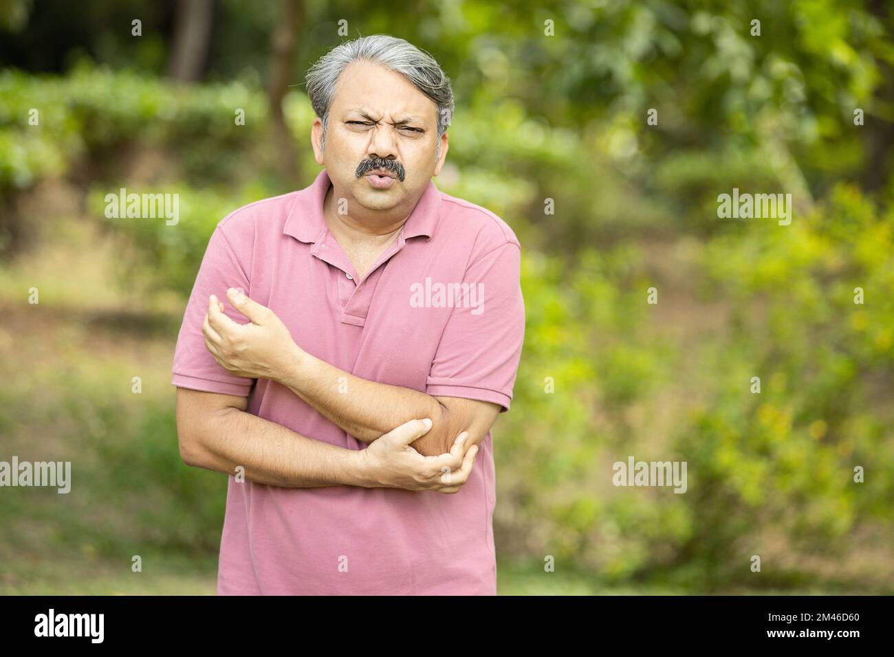 Indian senior man with arm pain standing outdoor at park. Asian Old man hand holding his elbow suffering from elbow pain. Health care. Stock Photo
