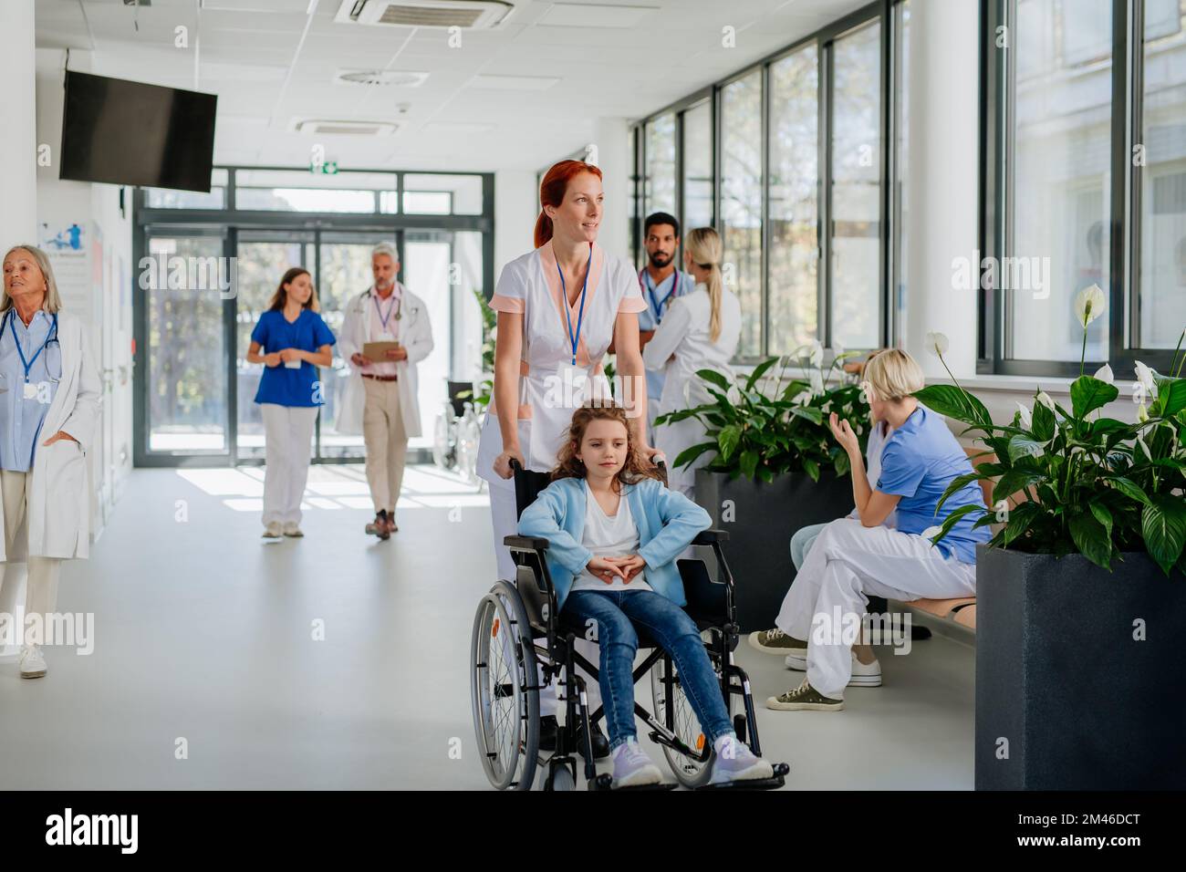 Young nurse pushing little girl on wheelchair at hospital corridor. Stock Photo