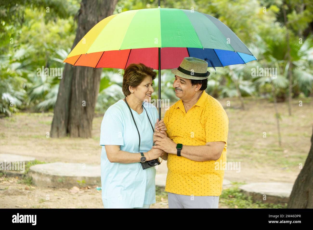 Happy indian senior couple standing under big colorful umbrella wearing casual cloths and hat outdoor at park, Retired old people enjoying holidays. Stock Photo