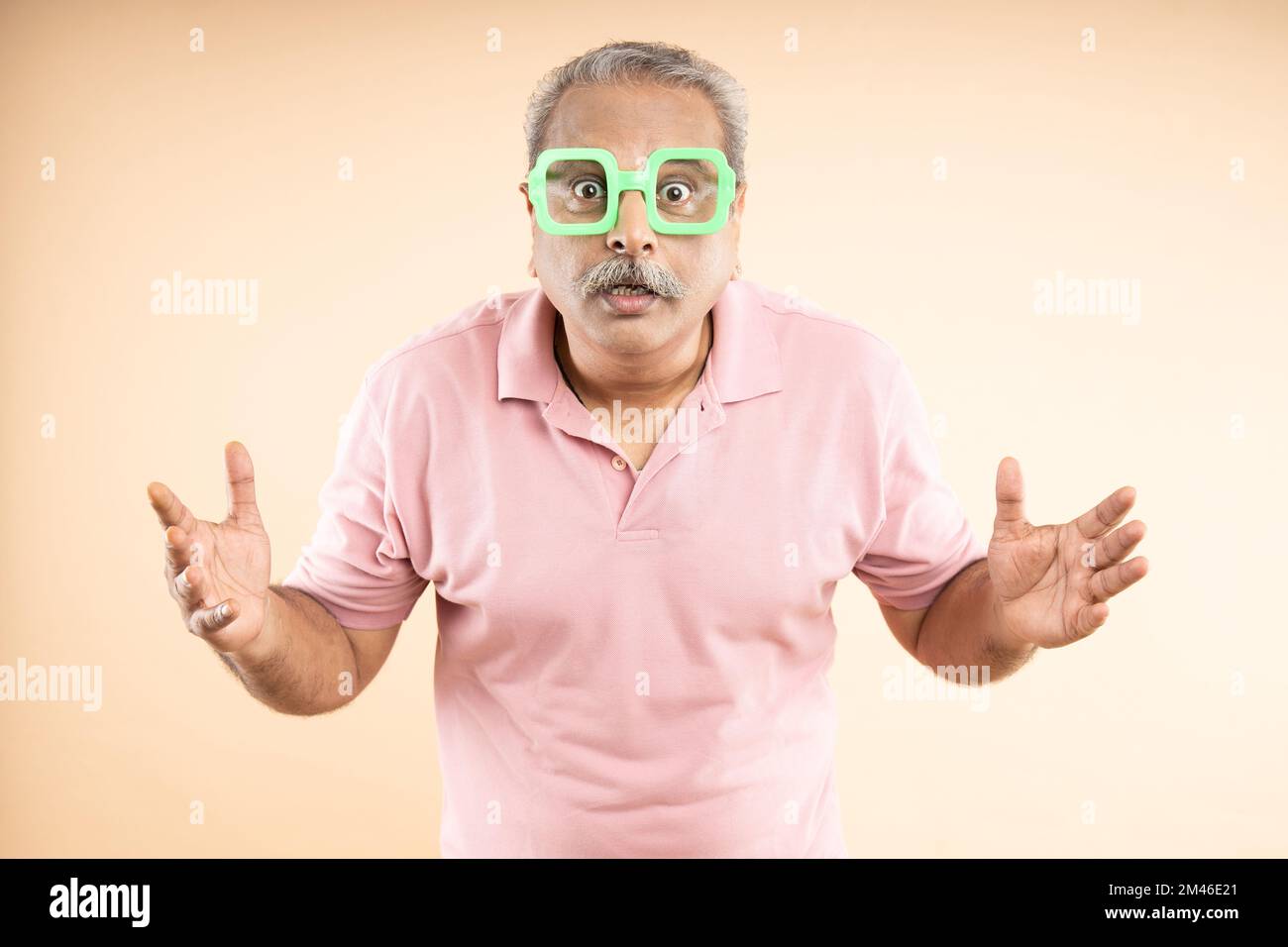 Amazed or surprised senior old indian man wearing funny sun glasses or eye wear standing isolated on beige background. Stock Photo