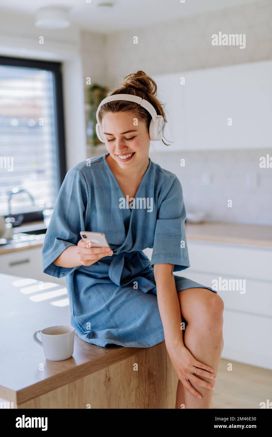 Young woman listening music, scrolling phone and enjoying cup of coffee at morning, in her kitchen. Stock Photo