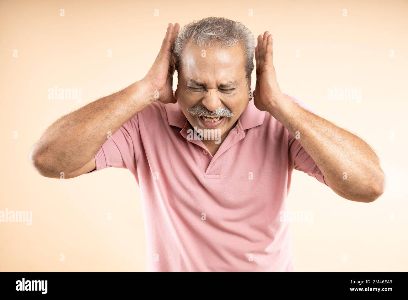 Irritated Indian senior man screaming with shut ears, disturbed by loud noise, standing over beige background. Stock Photo