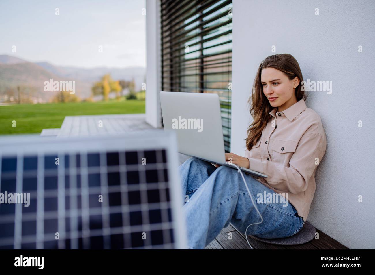 Young woman sitting on terrace, charging tablet trough solar panel. Stock Photo